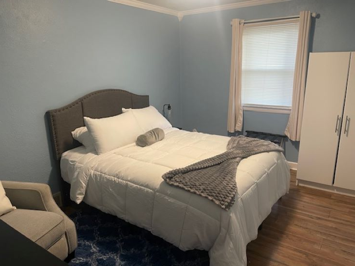 Bedroom bed with comforter, three pillow, and throw. also has a lamp and loveseat