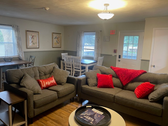 familyroom2.webp two gray couches with red and gray pillows. a coffee table with magazines and a dining room table with fours chairs (white/wood)